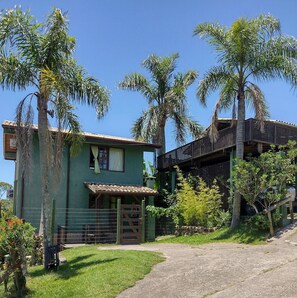 A two-story house with a green exterior, a balcony, and a fenced yard with palm trees. - Pousada Vale Verde Bungalows (Imbituba)