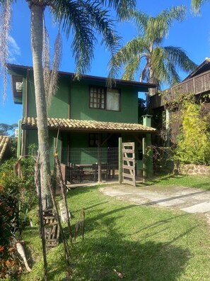 A green house with a tiled roof, a fenced yard, and palm trees. - Pousada Vale Verde Bungalows (Imbituba)