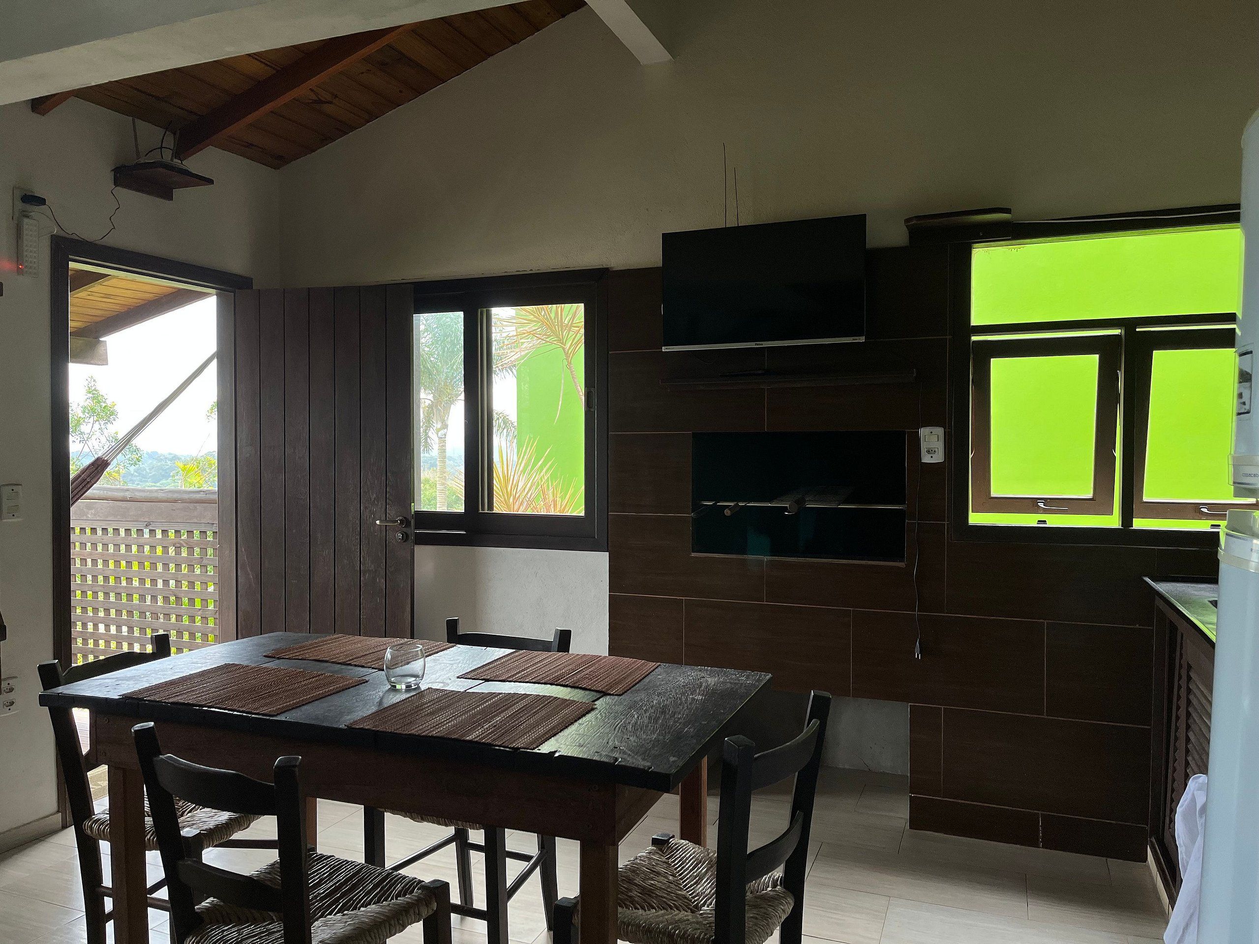 Uma área de refeições com mesa e cadeiras de madeira, janela com vista para palmeiras e TV embutida na parede.