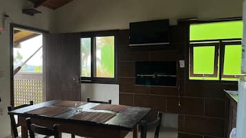 Uma área de refeições com mesa e cadeiras de madeira, janela com vista para palmeiras e TV embutida na parede.