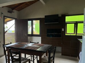 A dining area with a wooden table and chairs, a window with a view of palm trees, and a built-in TV on the wall. - Pousada Vale Verde Bungalows (Imbituba)