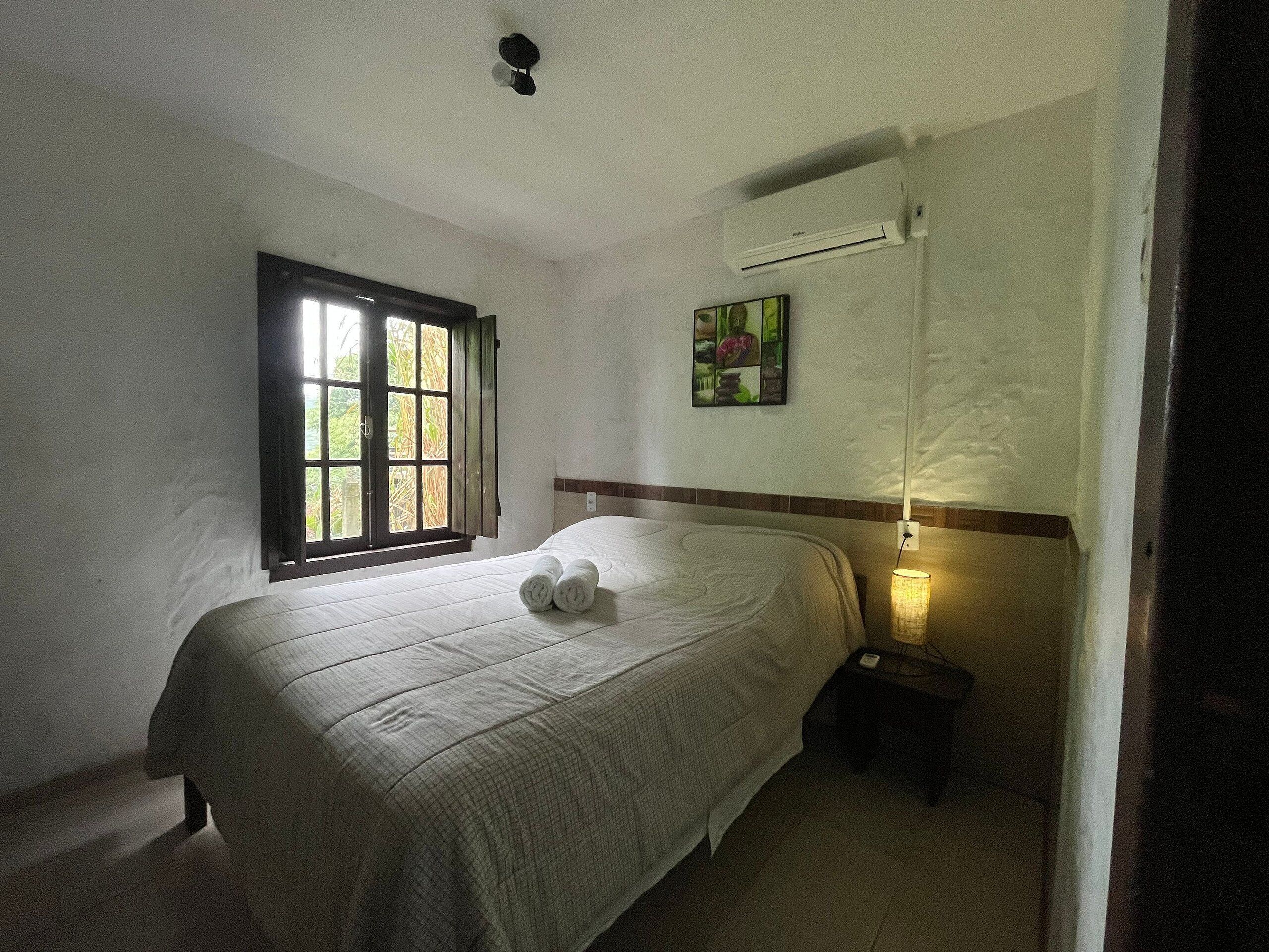 Cama bem arrumada com toalhas dobradas, janela com venezianas abertas, aparelho de ar condicionado e pequena mesa de cabeceira com abajur.