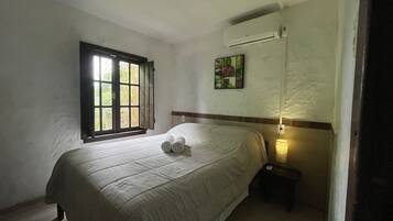 Cama bem arrumada com toalhas dobradas, janela com venezianas abertas, aparelho de ar condicionado e pequena mesa de cabeceira com abajur.
