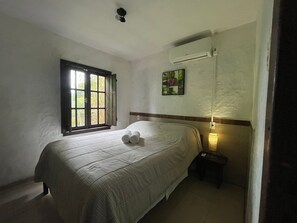 A neatly made bed with rolled towels, a window with open shutters, an air conditioning unit, and a small bedside table with a lamp. - Pousada Vale Verde Bungalows (Imbituba)