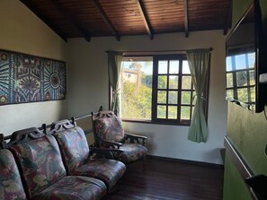 A room with a wooden ceiling, a large window with curtains, a sofa, and a wall with a decorative piece. - Pousada Vale Verde Bungalows (Imbituba)