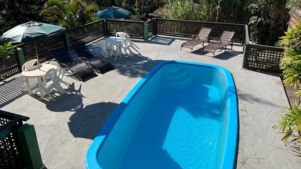 A rectangular swimming pool with lounge chairs and tables around it, surrounded by a fence and greenery. - Pousada Vale Verde Bungalows (Imbituba)