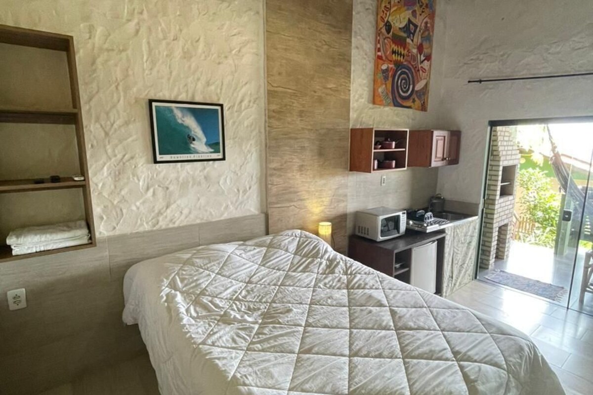 A bedroom with a bed, a shelf with towels, a painting, a microwave, and a view of the outdoors.