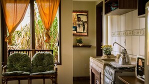 A room with a window overlooking a garden, a wooden chair, a kitchen with a stove and sink, and a wall with a framed picture. - Pousada Vale Verde Bungalows (Imbituba)
