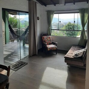 A room with a sofa, a chair, a window with a view of greenery, and a hammock outside. - Pousada Vale Verde Bungalows (Imbituba)