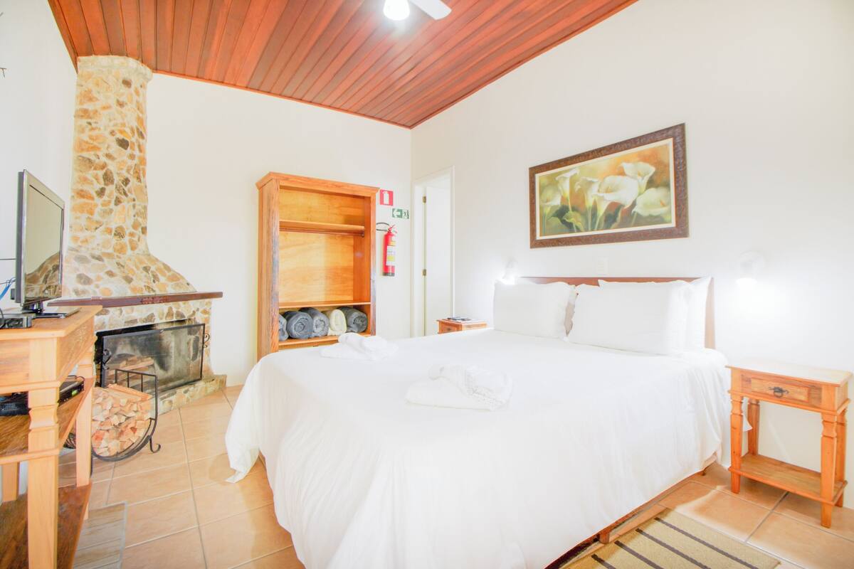 Habitación de hotel con chimenea de piedra, cama con ropa de cama blanca, cabecero de madera y un cuadro en la pared.