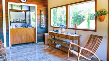 Um interior em madeira com uma mesa de recepção, uma cadeira, uma pequena mesa e uma janela com uma planta.