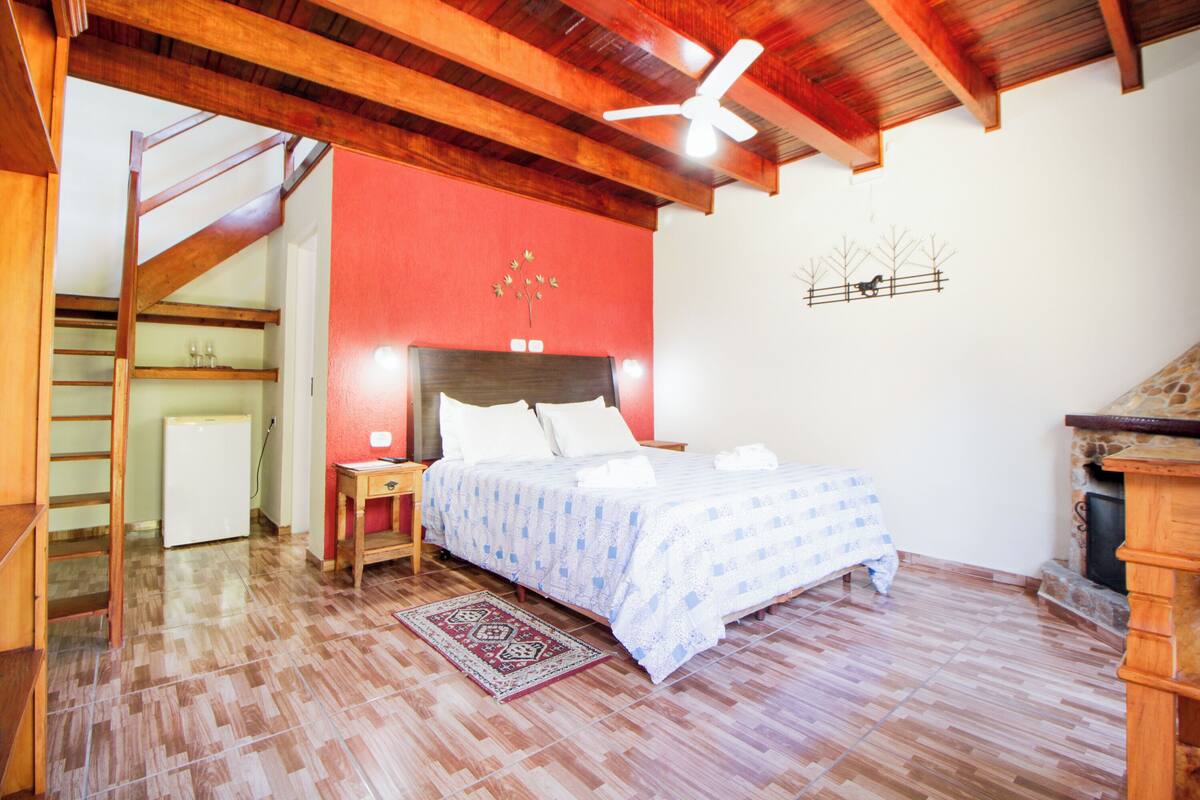 Un dormitorio con una cama, vigas de madera en el techo, un ventilador de techo, una mesita con lámpara, una pared roja y una chimenea.