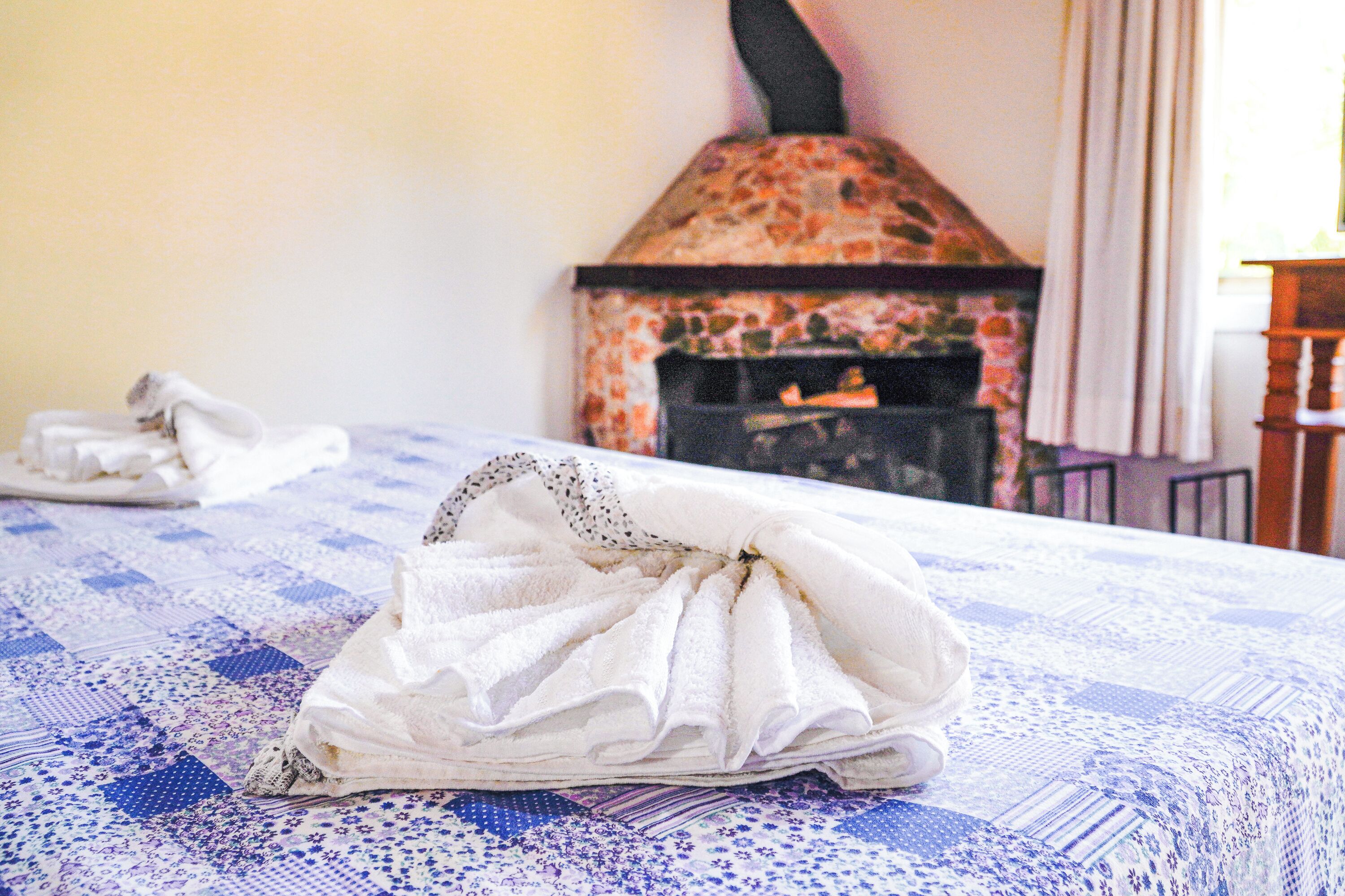 Uma cama com colcha xadrez azul e branca, uma toalha branca e uma lareira de pedra rústica ao fundo.
