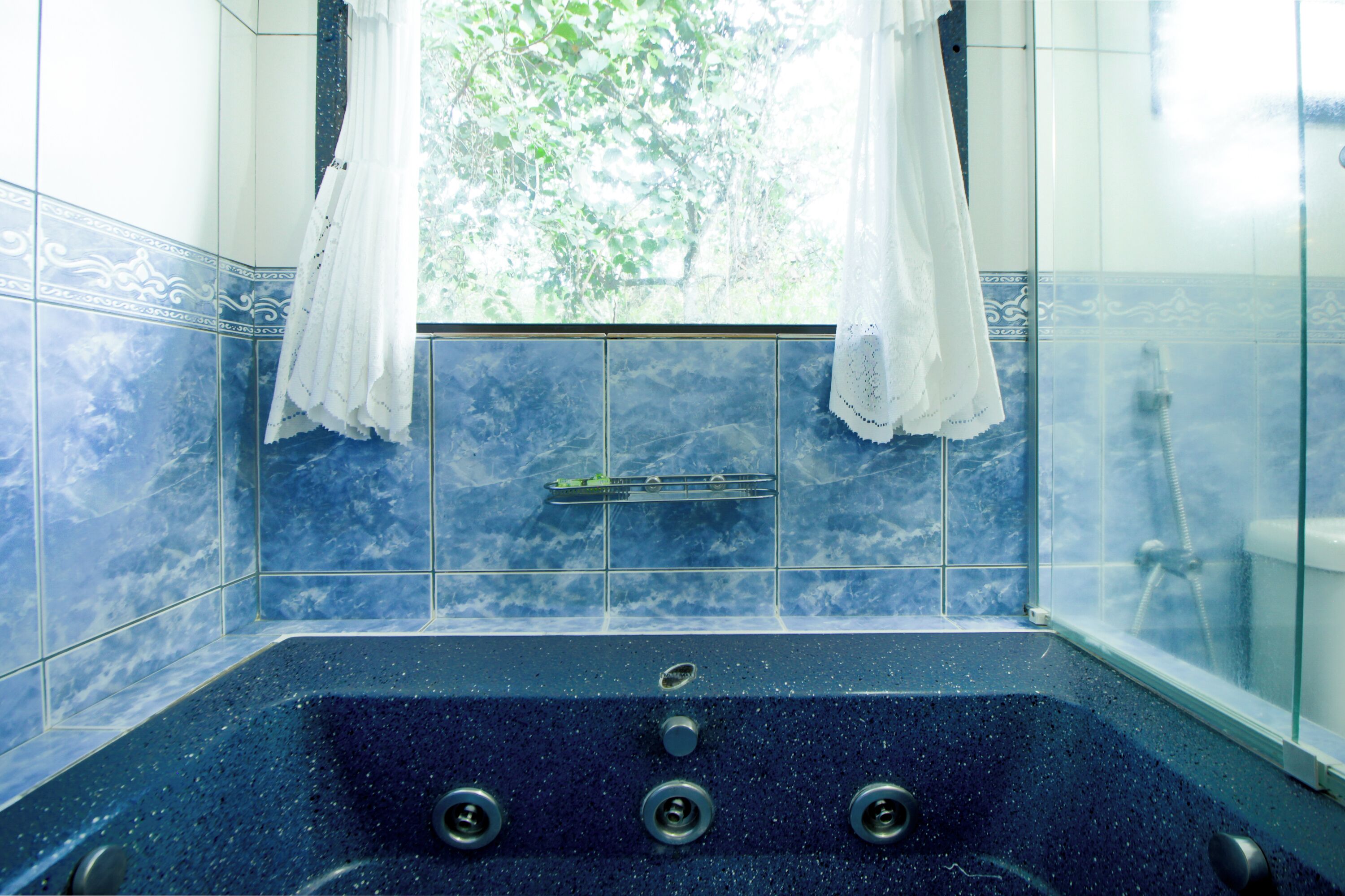 A bathroom with a large bathtub, a window with white curtains, and blue tiled walls.