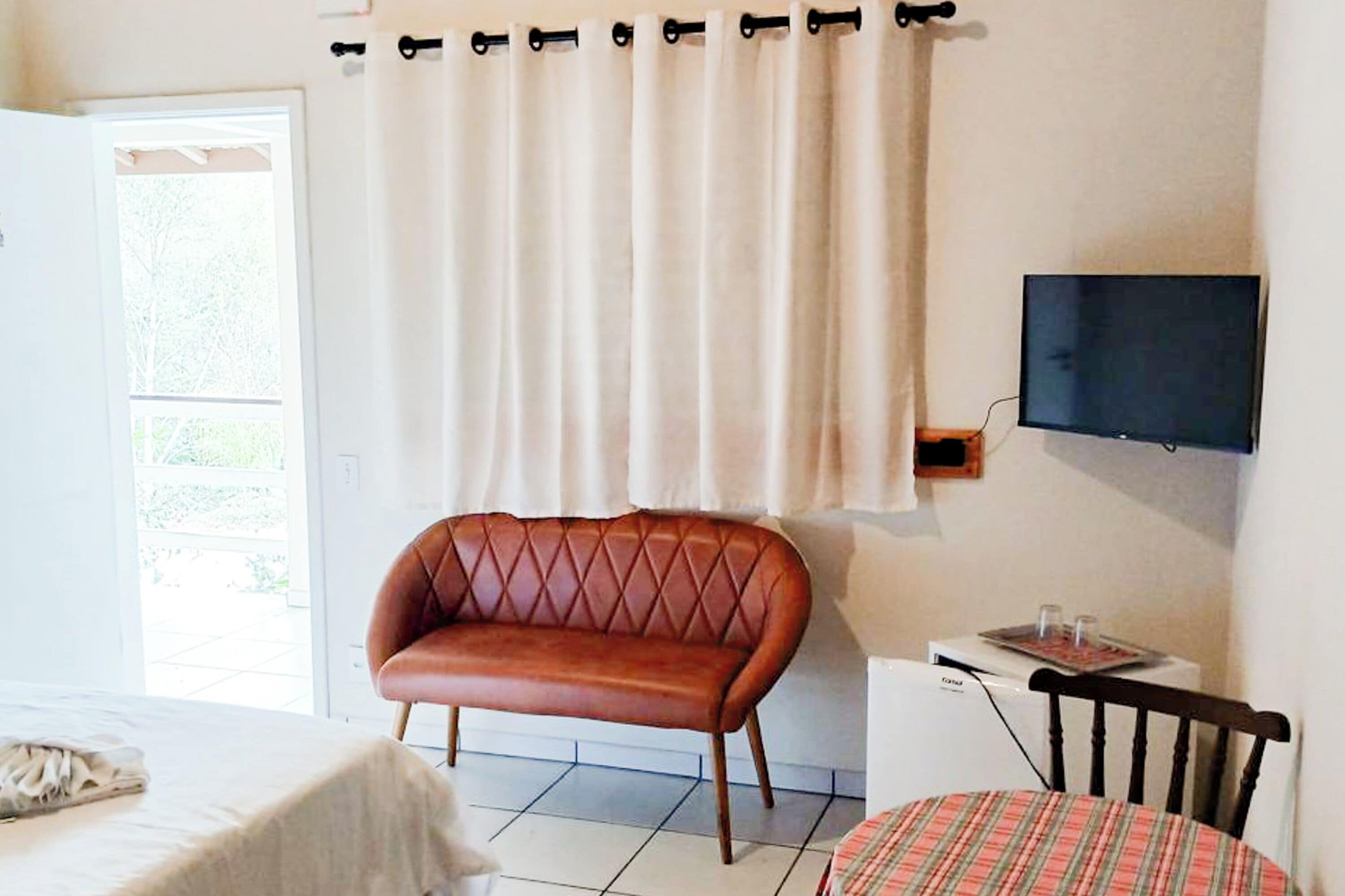 Une pièce comprenant un lit, un canapé en cuir marron, une petite table avec un téléviseur et une fenêtre avec des rideaux.