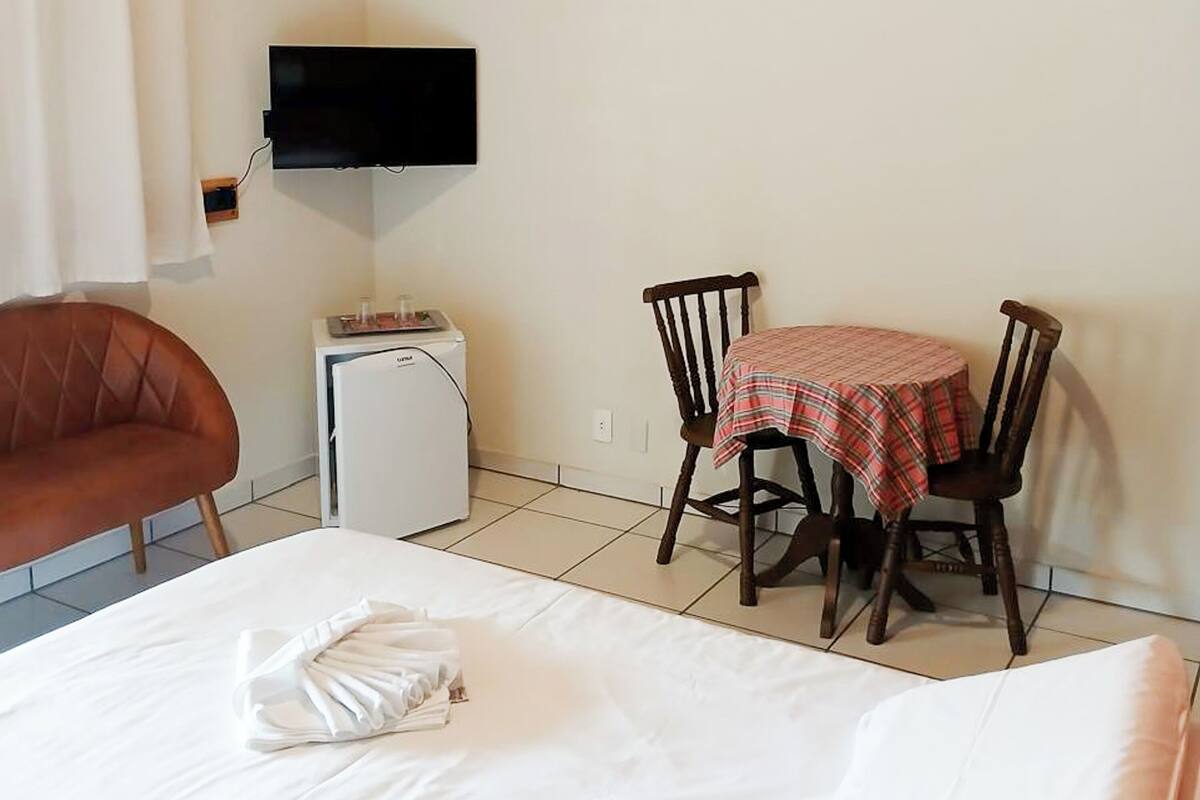 Habitación de hotel con cama, una mesita pequeña con mantel a cuadros, dos sillas de madera, televisor montado en la pared y un refrigerador blanco.
