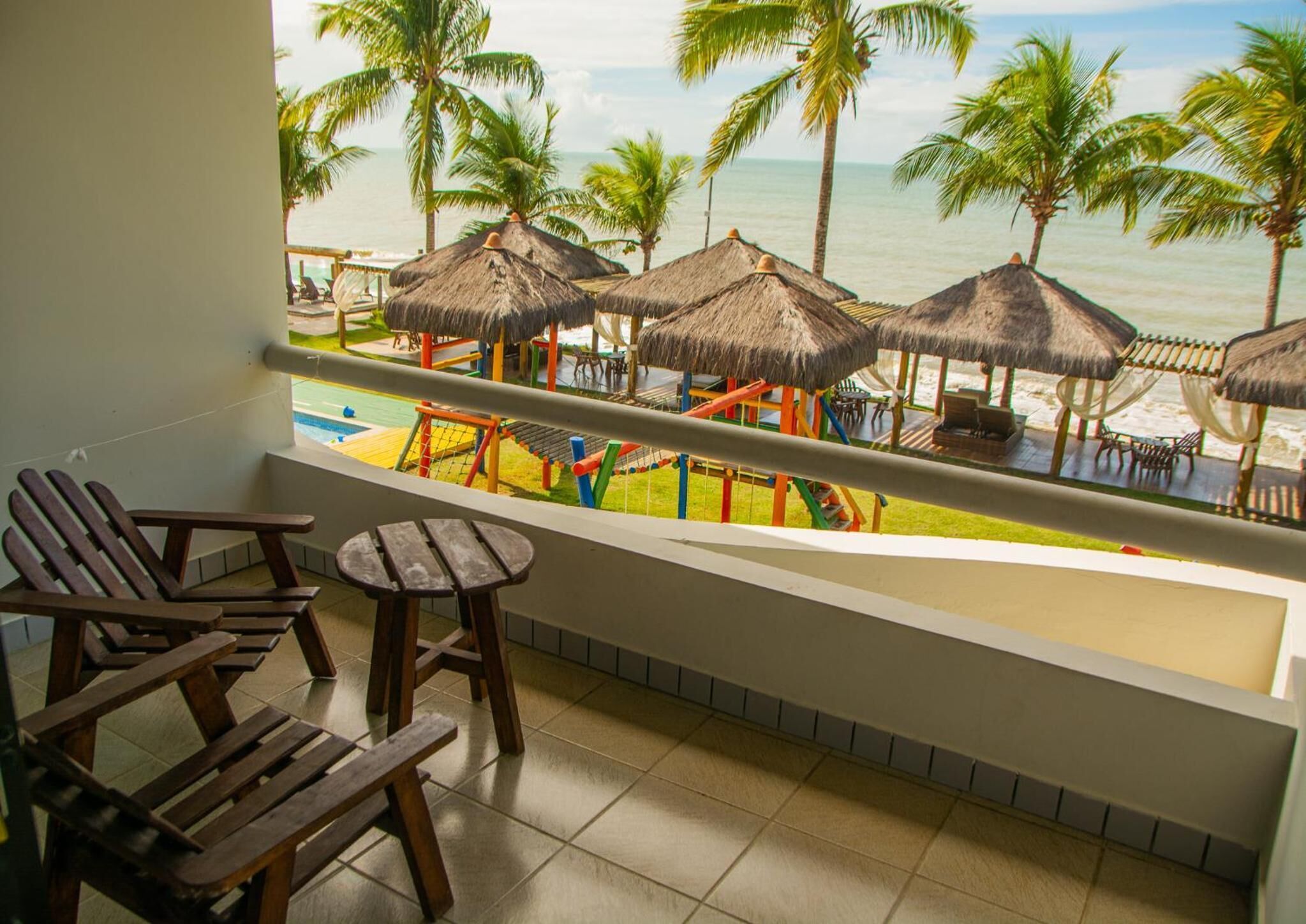A balcony with wooden chairs and a small table, overlooking a pool area with colorful play structures and palm trees.