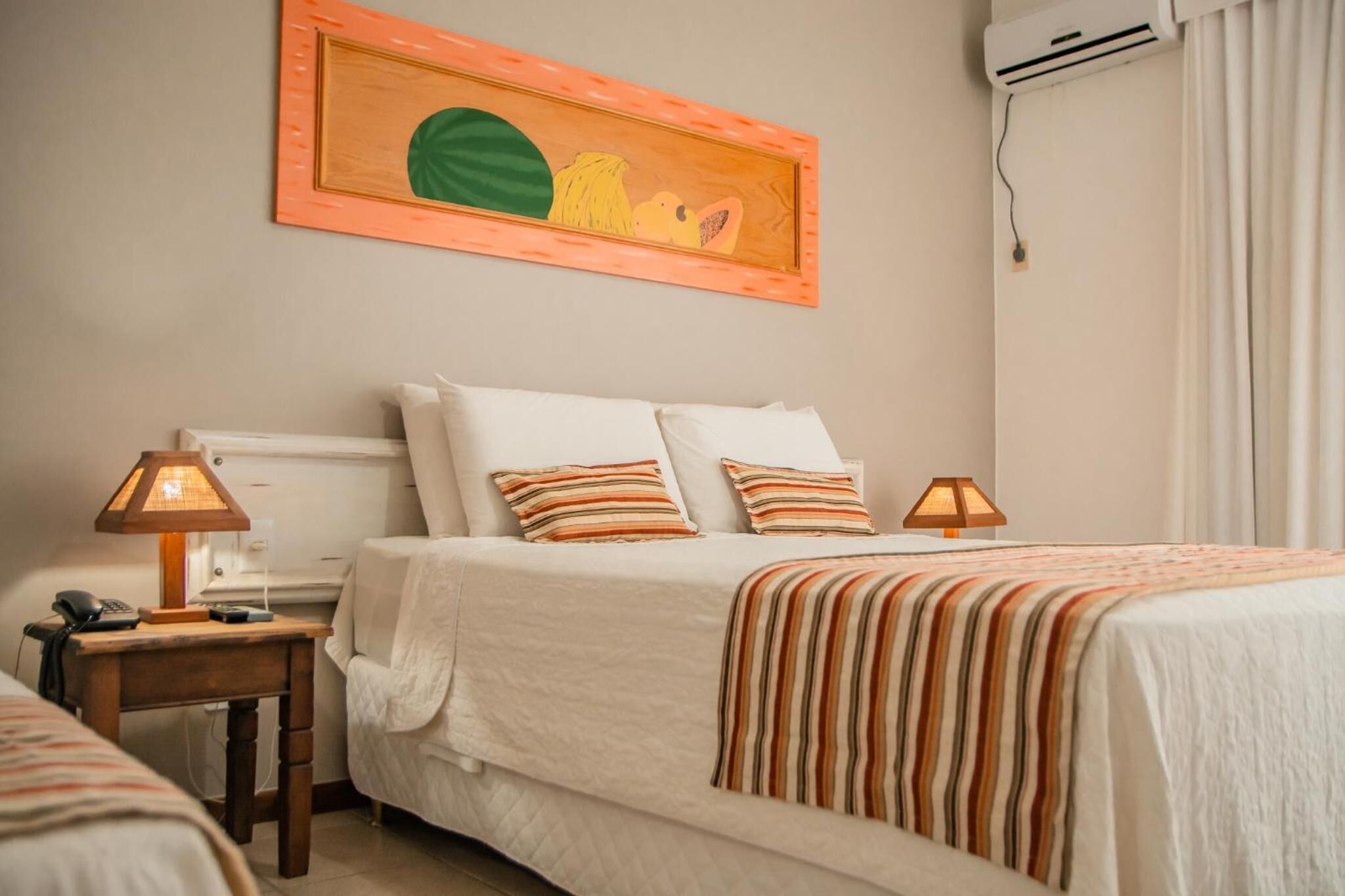 A hotel room with a bed, two bedside tables, a lamp, a painting, and an air conditioning unit.