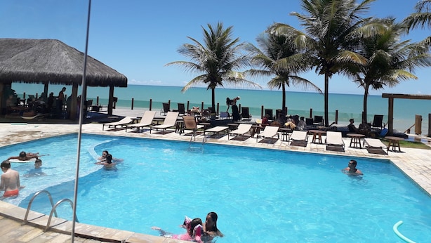 A pool area with lounge chairs and palm trees, surrounded by a wooden deck and a thatched-roof structure.