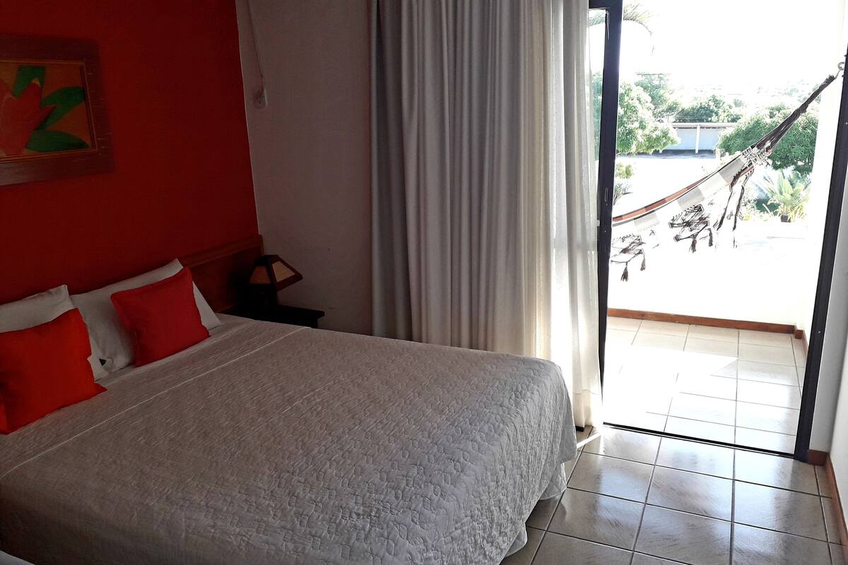 A bedroom with a bed, red pillows, a nightstand, a lamp, a window with curtains, and a view of a hammock outside.