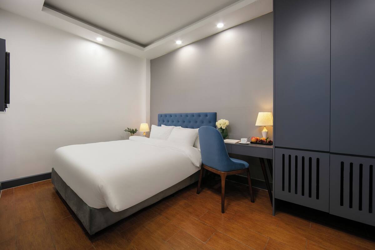 A hotel room with a bed, a desk with a chair, a lamp, and a wardrobe.