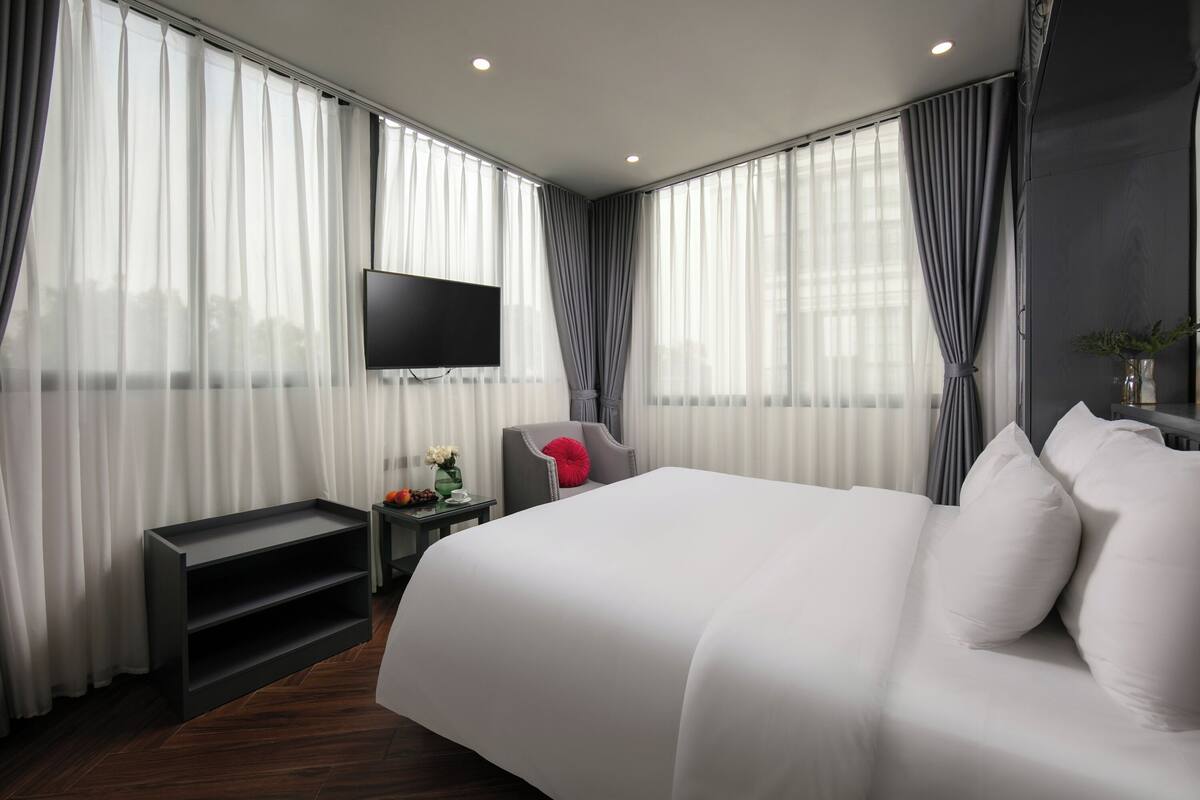 A modern hotel room with a large bed, a flat-screen TV mounted on the wall, a small side table with a vase of flowers, and a grey armchair.