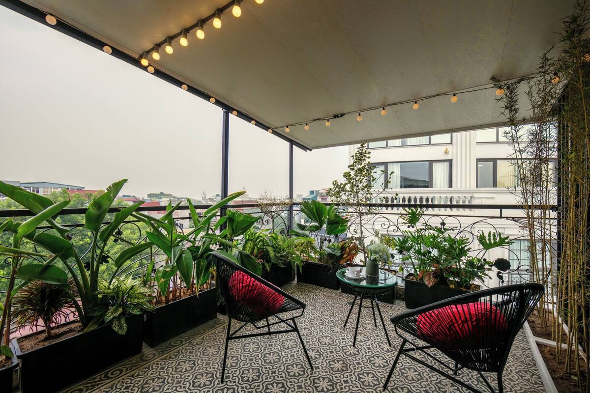A covered outdoor seating area with potted plants, two black chairs with red cushions, and a patterned floor.