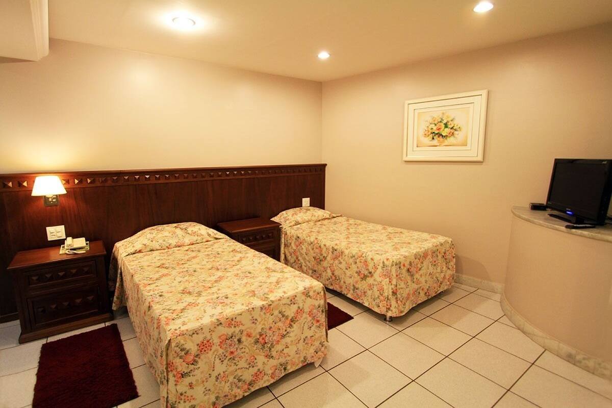 Une chambre d’hôtel avec deux lits simples, une tête de lit en bois, une table de chevet avec une lampe, un téléviseur posé sur un meuble et un tableau encadré au mur.