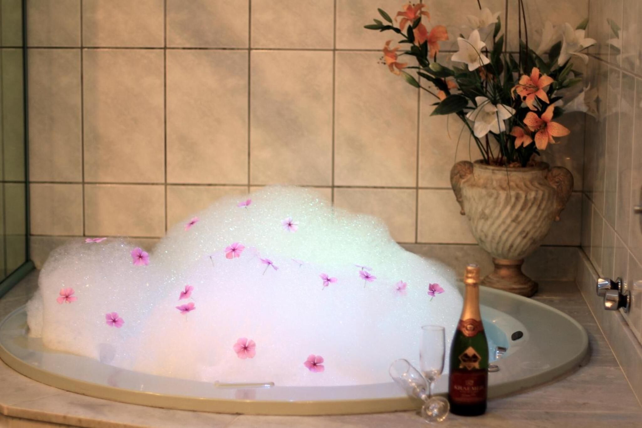 A bathtub filled with bubbles and pink flowers, a vase with flowers, and a bottle of champagne.