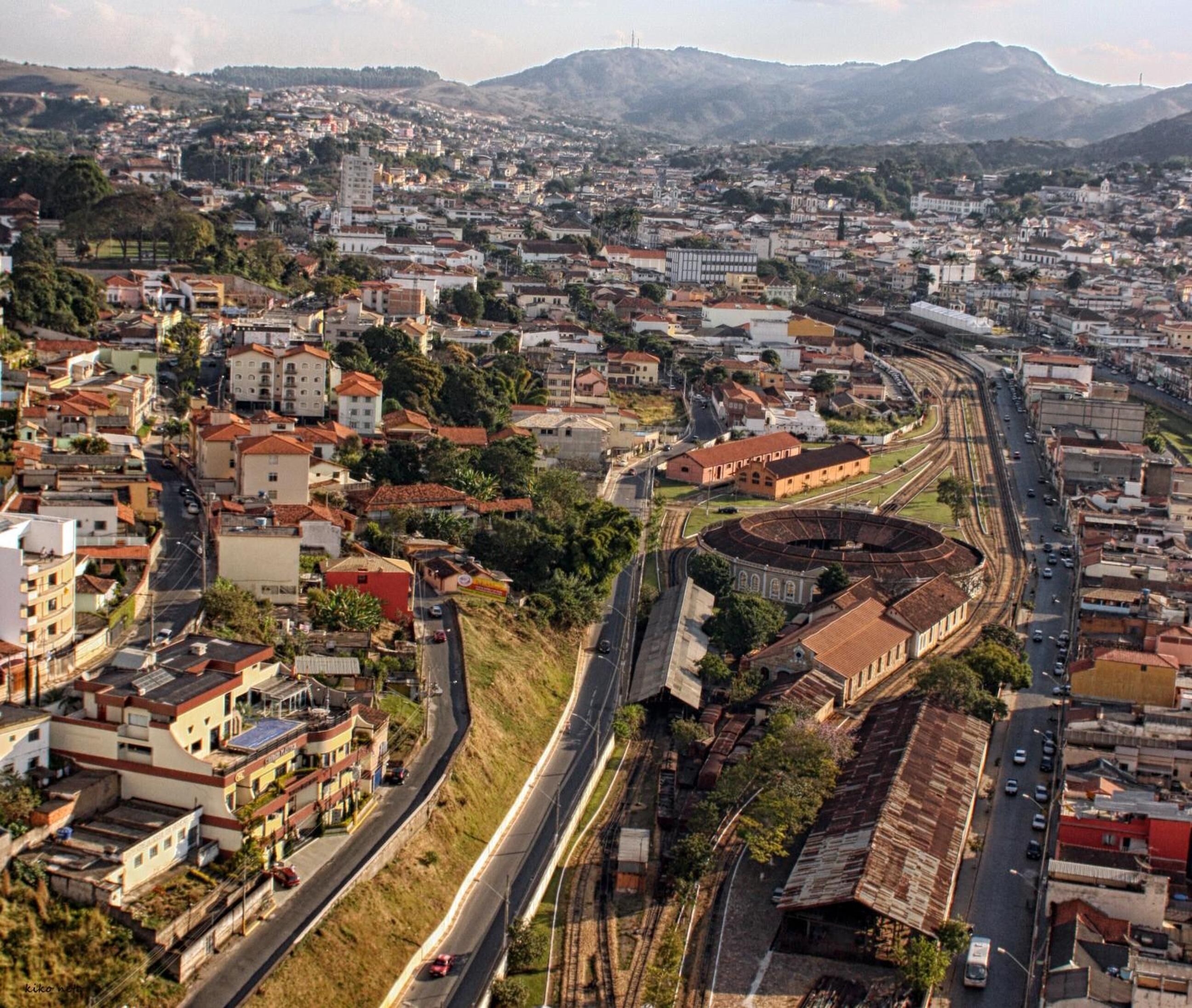 Vista aérea de uma cidade com uma estrutura circular imponente, possivelmente um estádio, rodeada por edifícios residenciais e estradas.