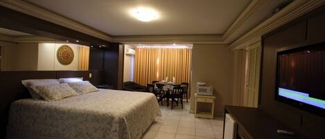 Habitación de hotel con cama, escritorio, televisión y zona de comedor con cortinas.