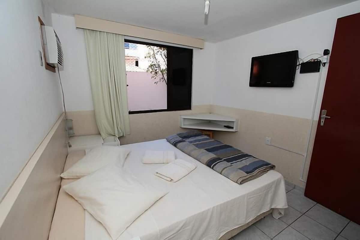 A small, clean room with a single bed, a window with a curtain, a mounted TV, and a door.