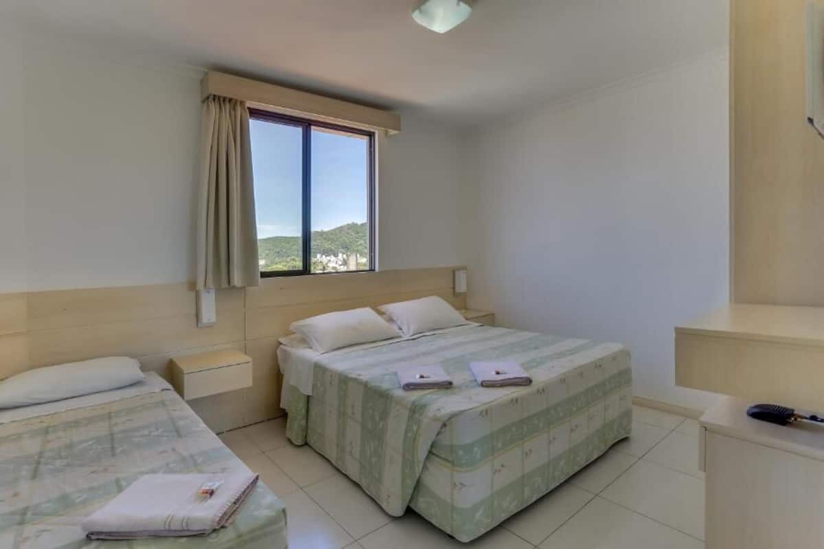 A bedroom with two beds, a window with a view, a bedside table, and a desk with a lamp.