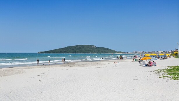 A beach with a sandy shore, umbrellas, and people. - Pousada Costão dos Ingleses (Florianopolis)