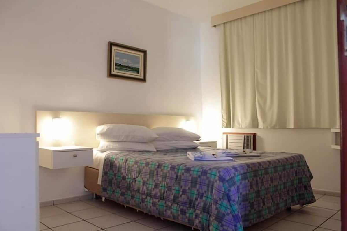 A bedroom with a bed, a bedside table, a nightstand, a framed picture on the wall, and a window with curtains.