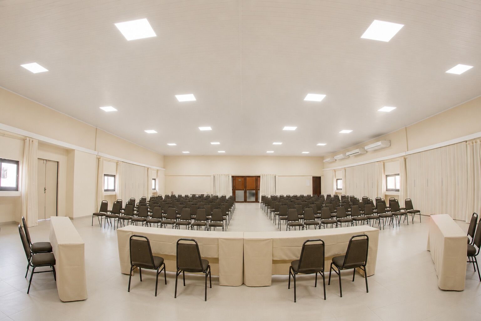 Un salón de conferencias con una larga mesa, varias filas de sillas y luces en el techo.