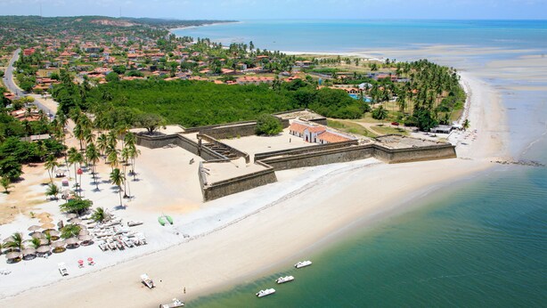 Vista aérea de una zona costera con playa, una estructura fortificada y un área residencial con edificios de techo rojo.