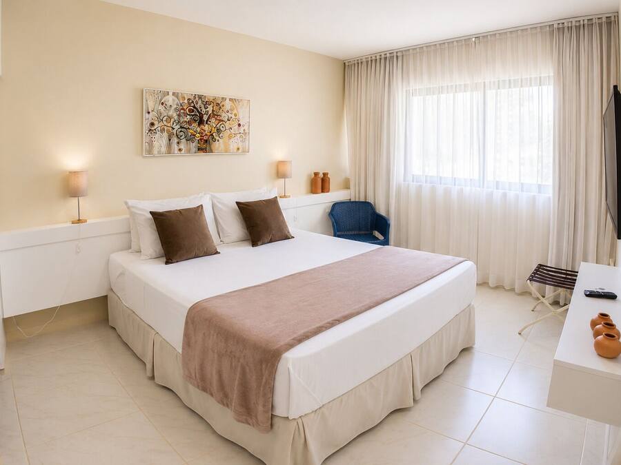 A hotel room with a bed, bedside lamps, a chair, a desk, a TV, and a large window with curtains.