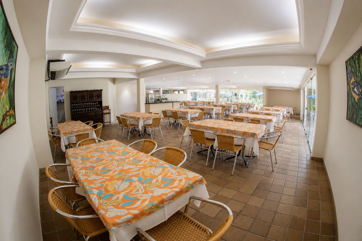 Uma sala de jantar com várias mesas e cadeiras, uma cozinha ao fundo, e vista através de janelas amplas.