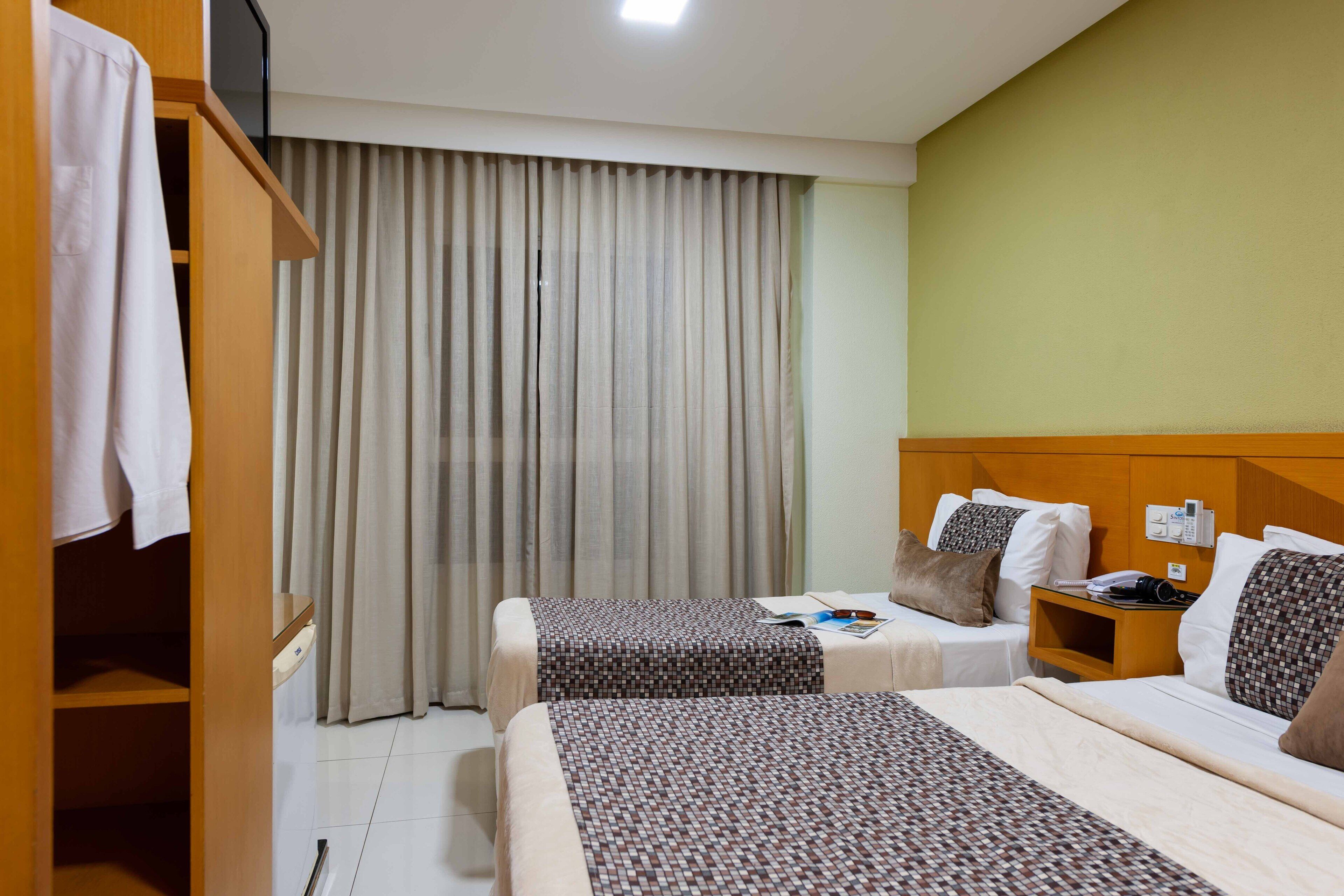 A hotel room with two beds, a wooden wardrobe, a nightstand with a lamp, and a window with curtains.