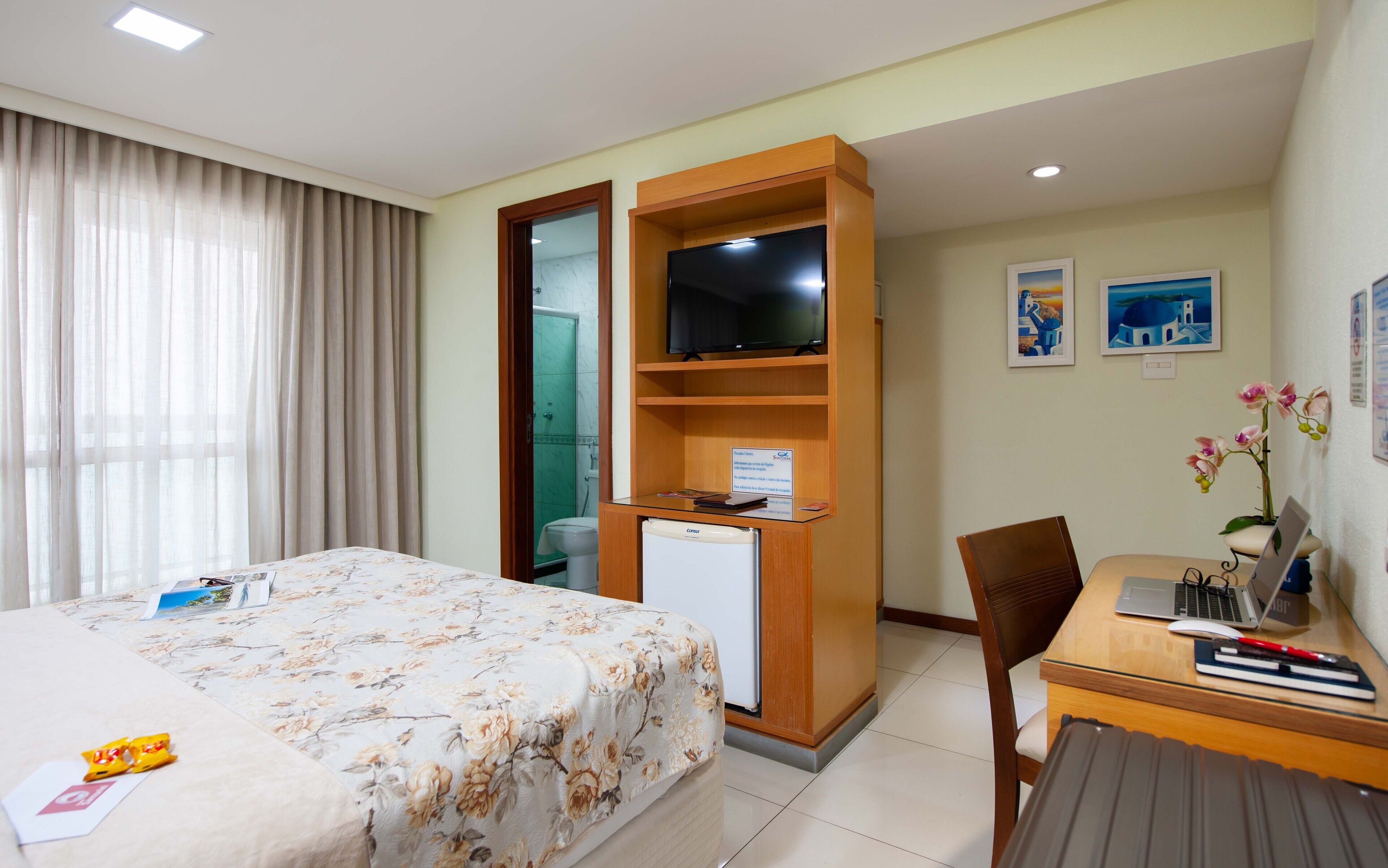 Habitación de hotel con cama, escritorio con laptop, refrigerador pequeño, televisor en estante de madera y zona de ducha.