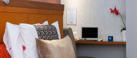 A hotel room with a bed, a desk with a laptop, a small potted plant, and a framed picture on the wall.