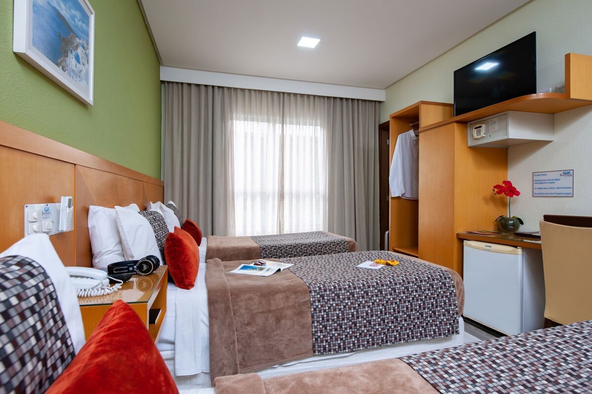 A hotel room with two beds, a desk, a TV, a minibar, and a window with curtains.