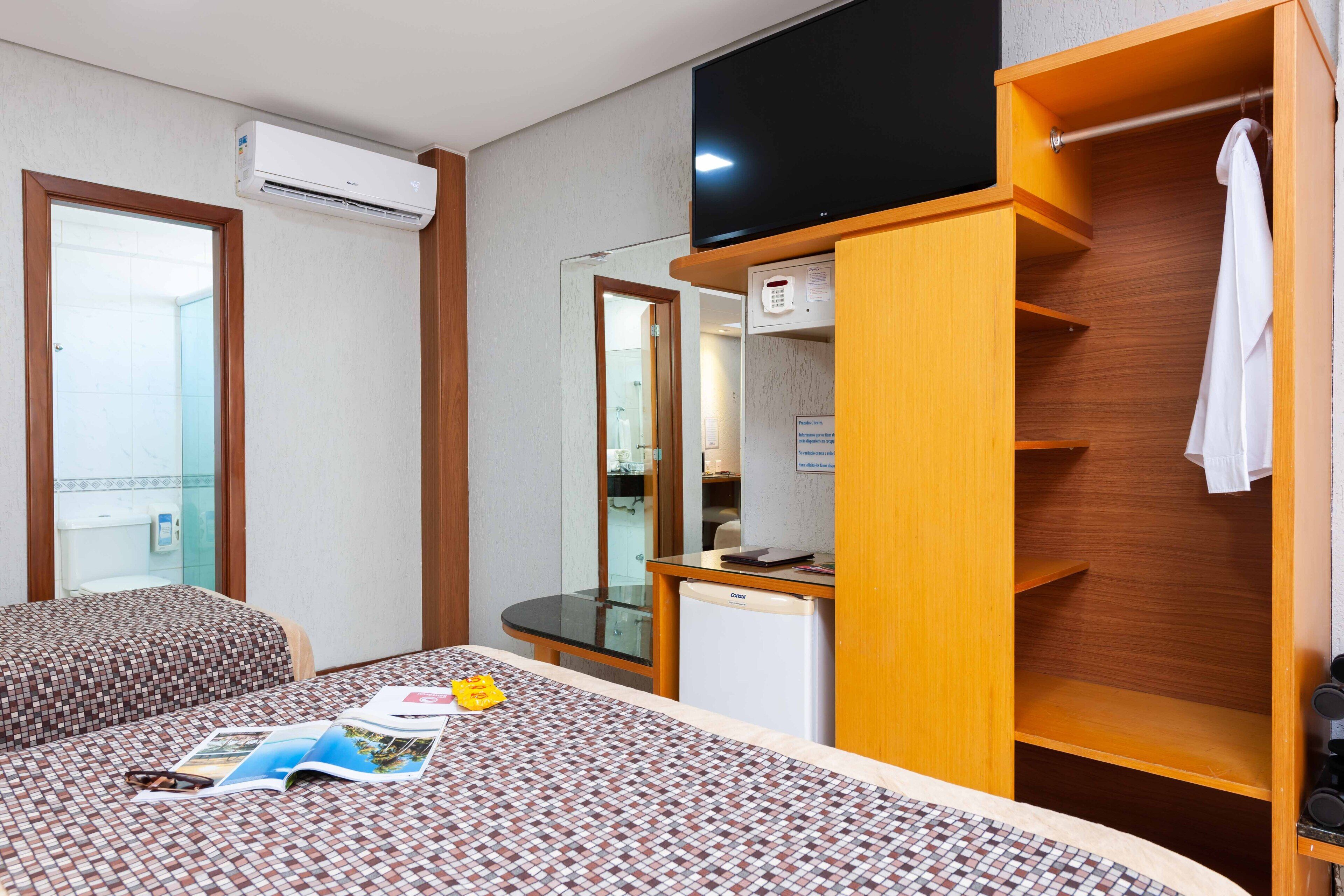 A hotel room with a bed, a small desk, a television, an air conditioning unit, and a wardrobe.