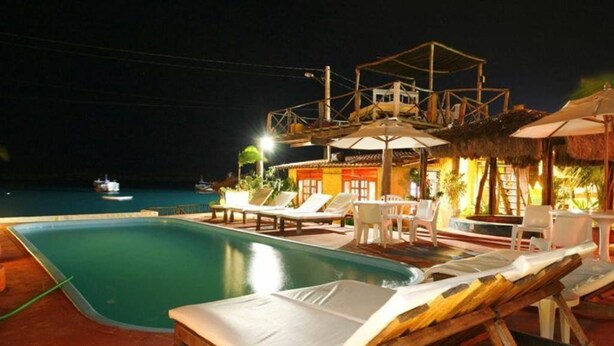 A pool area with lounge chairs and umbrellas, a wooden deck, and a building with a thatched roof. - Chalé Oásis (Galinhos)