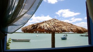 A view through a window with a thatched umbrella, a boat, and calm water. - Chalé Oásis (Galinhos)
