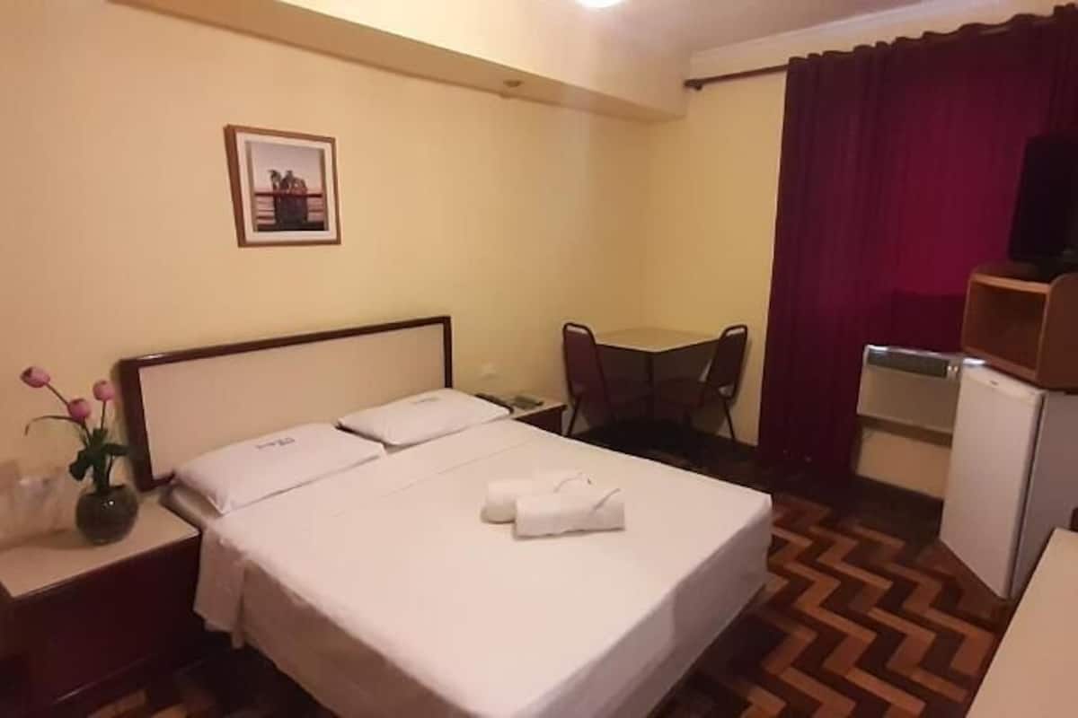 Habitación de hotel con cama, escritorio, silla, televisor y un refrigerador pequeño.