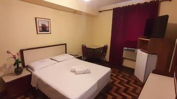 A hotel room with a bed, a desk, a chair, a TV, and a small refrigerator.