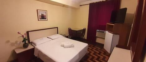 A hotel room with a bed, a desk, a chair, a TV, and a small refrigerator.