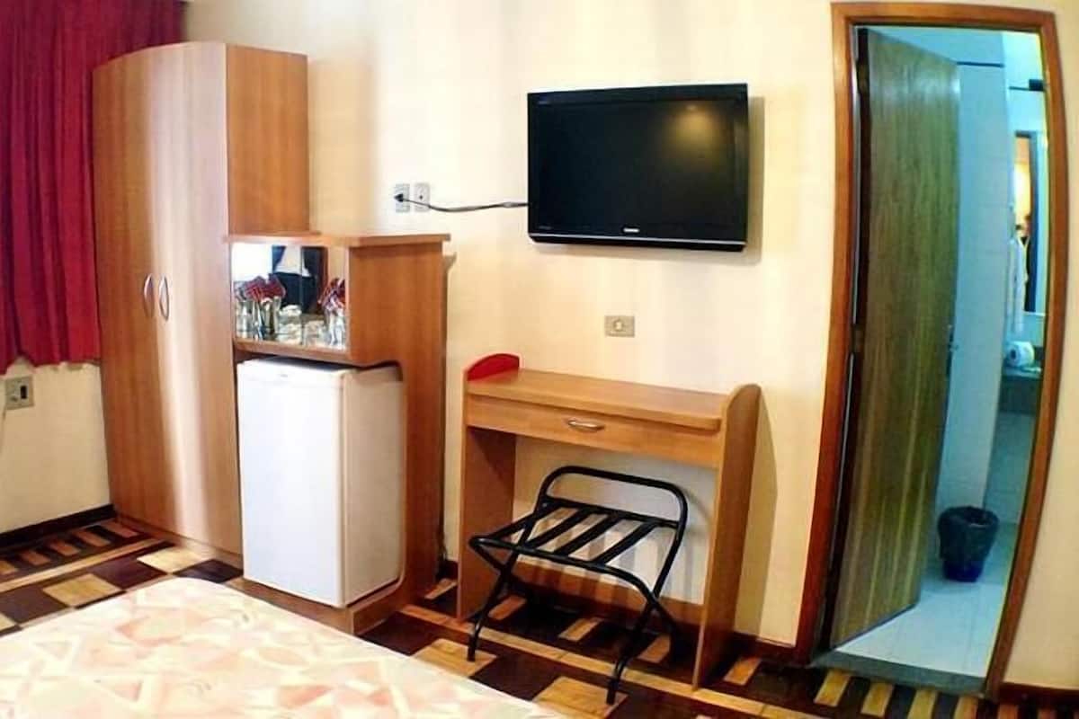 Una habitación de hotel compacta con cama, escritorio, silla, televisor, refrigerador y cortina roja.